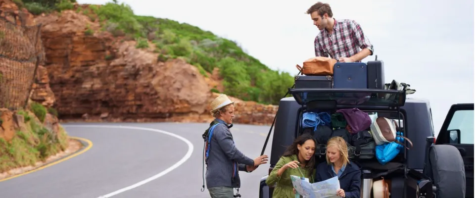 Group of friends checking a map beside their luggage-loaded vehicle on a scenic coastal road, practicing spring break safety by planning their travel route together.