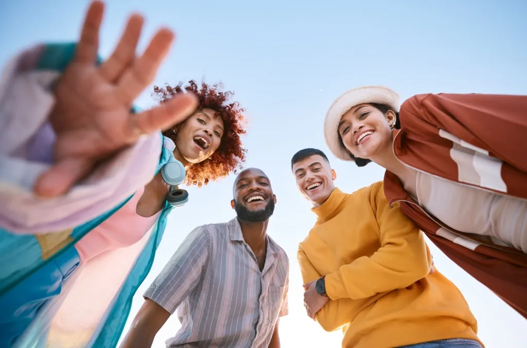 Four smiling friends looking down at the camera against a clear blue sky, enjoying spring break safely while spending time outdoors as a group.