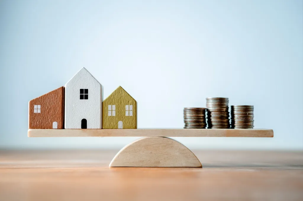 Three miniature wooden houses and stacks of coins balanced on a wooden seesaw, visually illustrating how insurance rates are determined based on risk assessment.