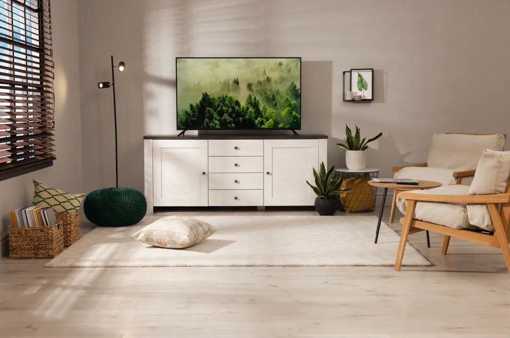 Bright, modern living room with natural sunlight streaming through wooden blinds, cozy seating, indoor plants, and a TV displaying a lush forest scene, as a background for summer safety tips to keep an apartment cool, secured, and comfortable.