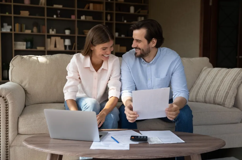 A couple sits together on a beige sofa, reviewing documents and using a laptop with bills and a calculator on the coffee table, thoughtfully discussing how much renters insurance they need.