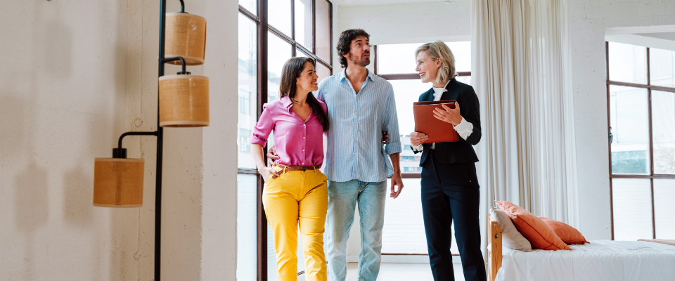 A real estate agent holding a clipboard showing a bright, modern apartment to a young couple during a rental walkthrough, illustrating the evaluation step in how to get approved for an apartment.
