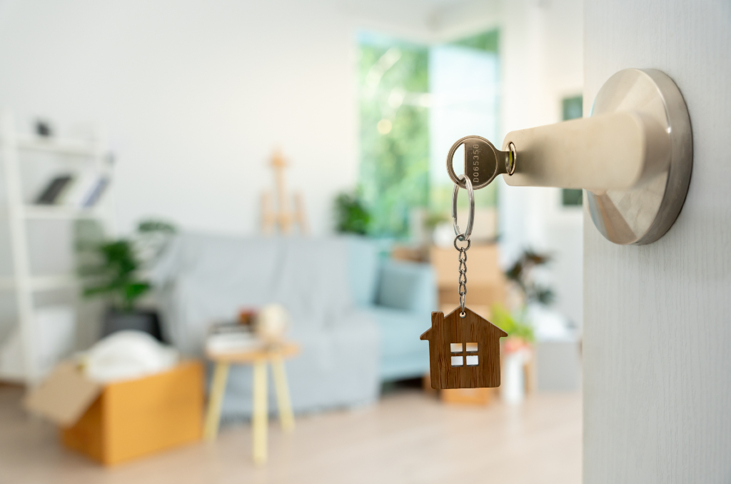 A key with a wooden house keychain inserted into an apartment door lock, with moving boxes and a furnished living room visible in the background, representing how to get approved for an apartment.