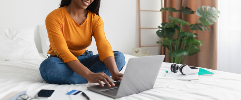 A smiling woman sitting cross-legged on a bed using a laptop with a credit card nearby, researching how to get approved for an apartment rental application online.