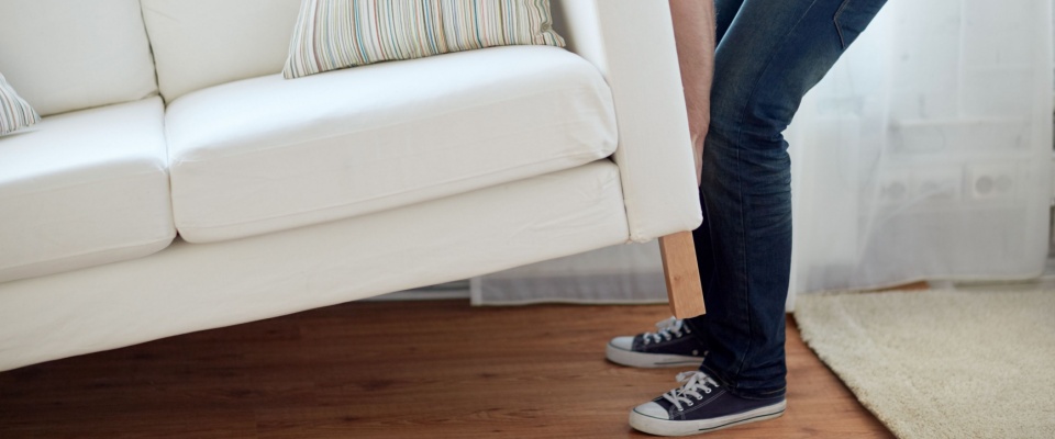 Person lifting a white sofa with striped pillows in a bright living room, for furniture rearrangement and fresh interior updates, as part of spring home improvements.