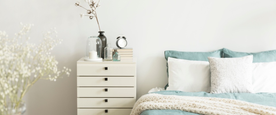 Bright bedroom showcasing spring home improvements with fresh bedding, decorative pillows, a cozy knit blanket, and a modern nightstand with vases, books, and an alarm clock.