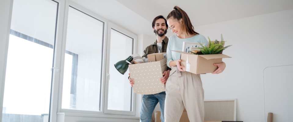A couple moving in together, carrying moving boxes filled with household items and plants in a bright, unfurnished apartment, highlighting the need to update a renters insurance policy when sharing a flat.