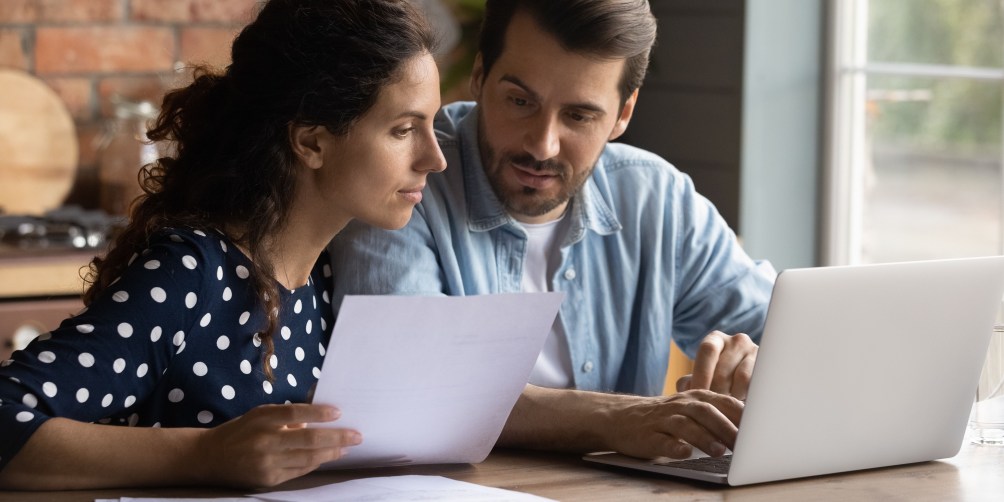 Couple sitting at a kitchen table reviewing paperwork and using a laptop as part of the process to transfer renters insurance.