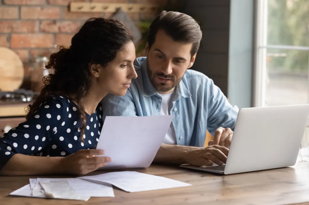 Couple sitting at a kitchen table reviewing paperwork and using a laptop as part of the process to transfer renters insurance.