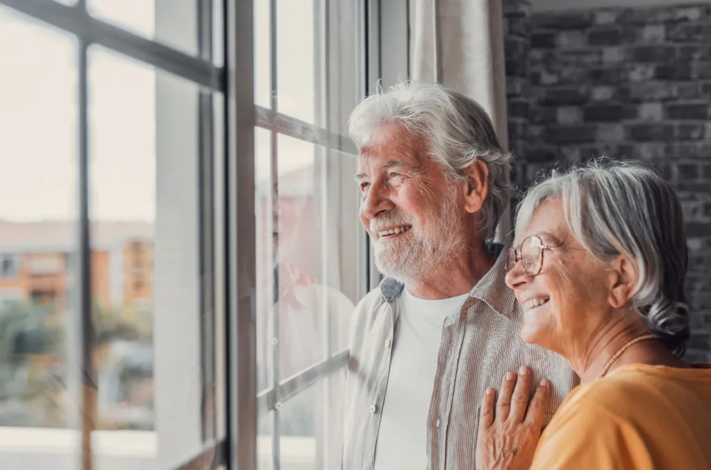 An senior couple stands together by a window in a cozy apartment, quietly looking outside and reflecting on the implications of downsizing.