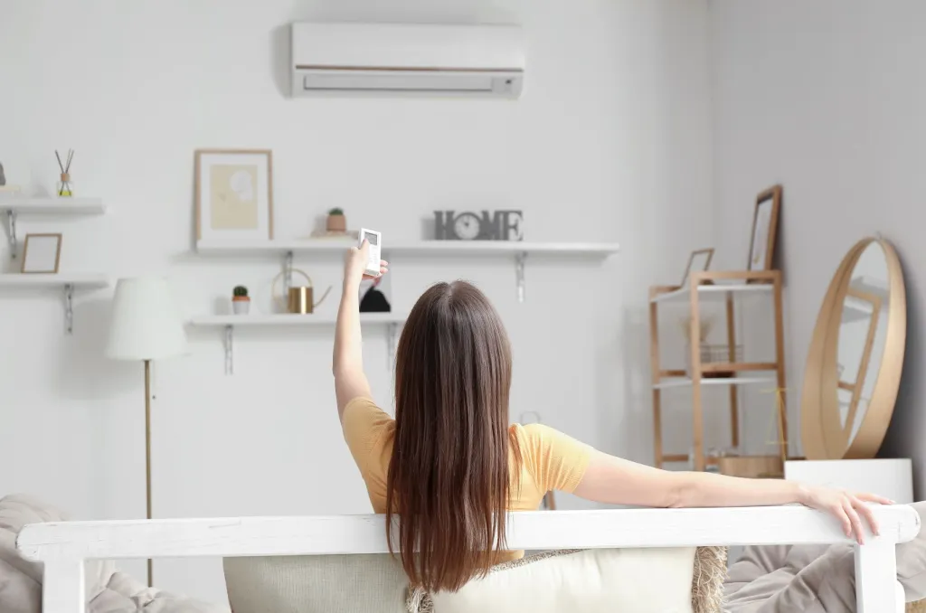 Woman sitting on a sofa in a bright living room, using a remote to adjust the air conditioner, illustrating how to lower the electric bill in summer by optimizing thermostat settings.