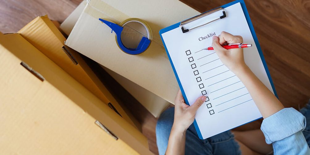 A person sitting on a wooden floor with a checklist on a clipboard, writing with a red pen, surrounded by cardboard boxes and packing tape.