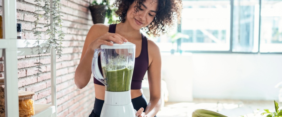 Woman in a sunlit apartment kitchen blending a green smoothie, illustrating how to prepare your apartment for summer by purchasing appliances perfect for the warm season and by maximizing natural light.