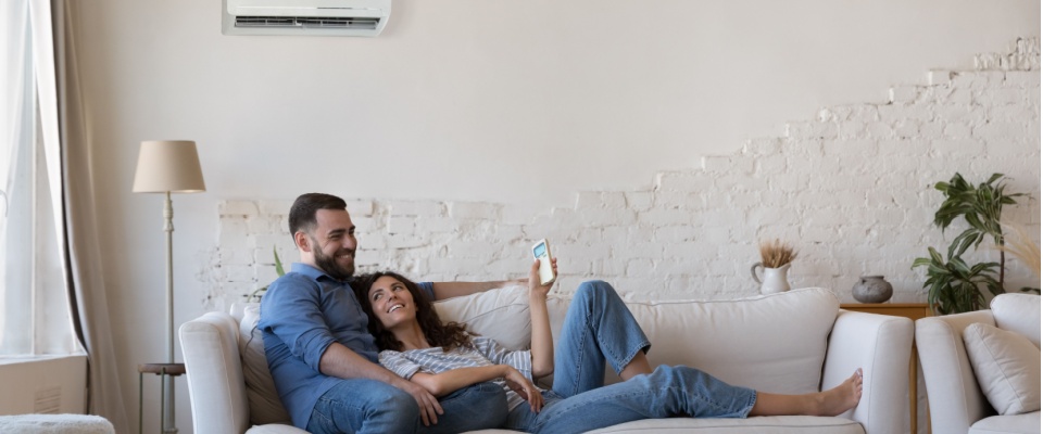 Couple relaxing on a white sofa in a bright living room with an air conditioner overhead, illustrating how to prepare your apartment for summer by creating a comfortable space with efficient climate control.