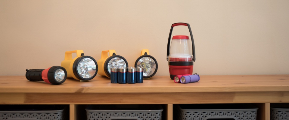 Multiple flashlights, lanterns, and a selection of extra batteries neatly arranged on a wooden shelf, illustrating essential supplies to include in a power outage emergency kit.