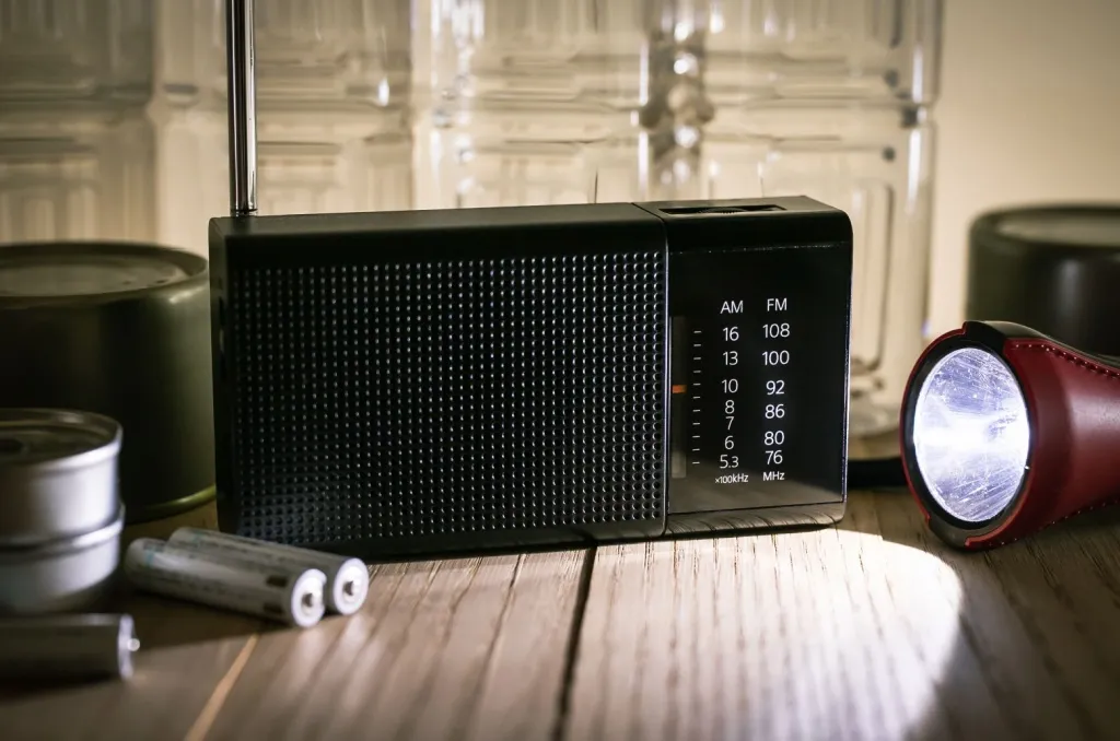 Battery-powered radio, flashlight, and extra batteries displayed on a wooden table, demonstrating essential items to include in a power outage emergency kit.