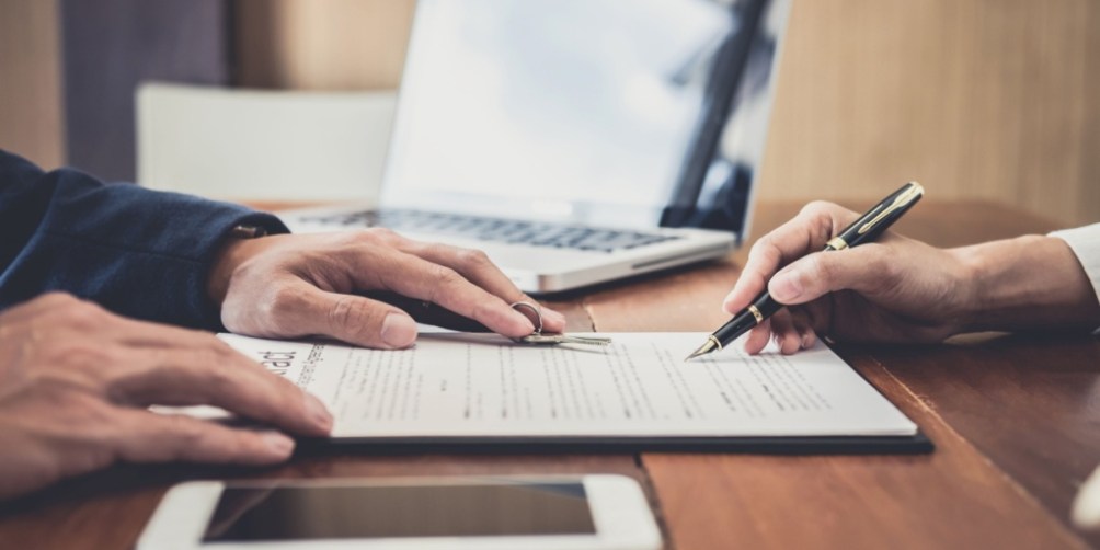 Two people signing a lease agreement at a wooden table with a laptop and tablet nearby, after reviewing important questions to ask before signing a lease.