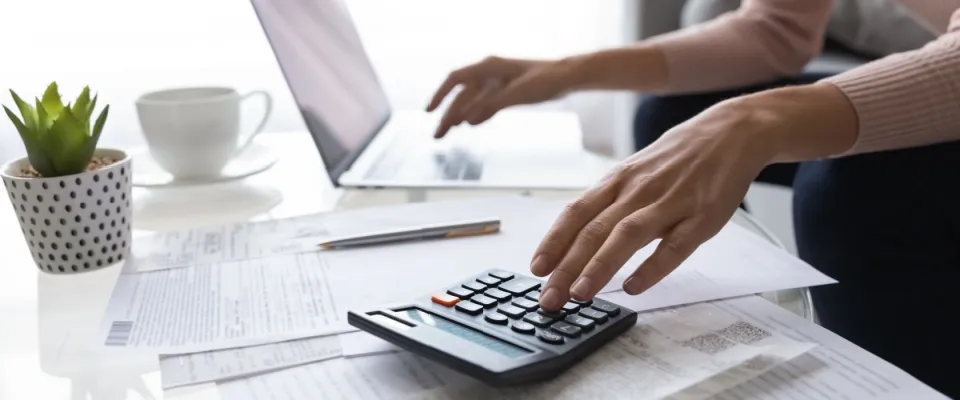 Person in front of a white coffee table, with a laptop, calculator, apartment lease agreement, a potted plant and a white cup, calculating rent costs. 