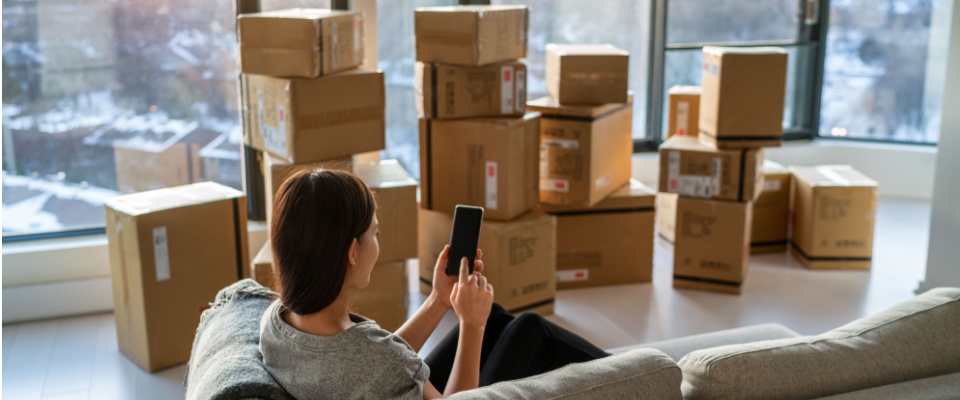 Woman sitting on a couch using a smartphone, surrounded by moving boxes in a modern apartment, considering transferring renters insurance instead of cancelling it.