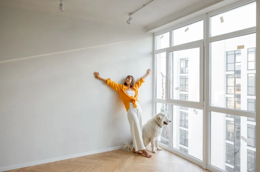Bright modern apartment interior with white walls, featuring a person standing near a large window alongside a large white dog, feeling joyful after successfully renting with pets.