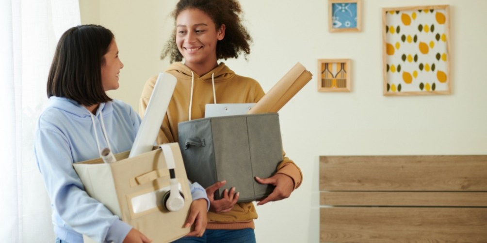 Two roommates standing in a bright bedroom, each holding boxes filled with personal belongings, possibly talking about renters insurance with roommates.