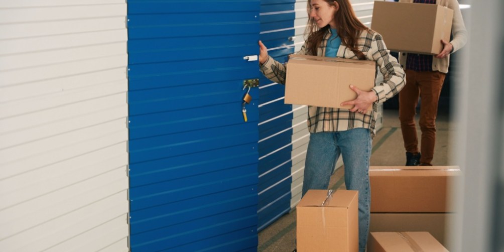Two people carrying cardboard boxes and unlocking a blue storage unit, visually illustrating how renters insurance covers storage units by protecting personal belongings stored off-site.