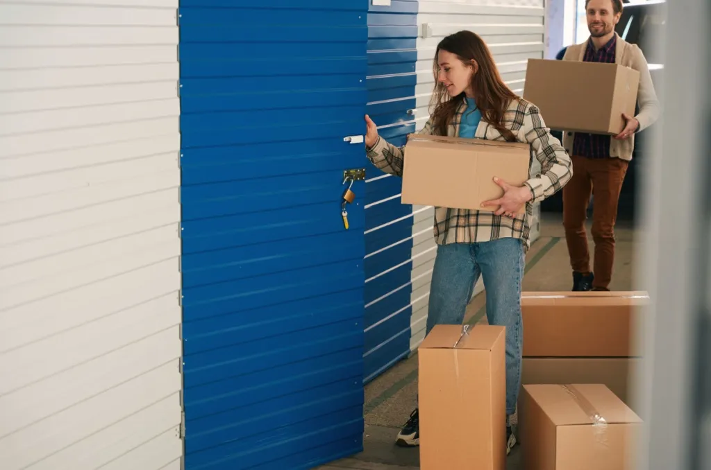 Two people carrying cardboard boxes and unlocking a blue storage unit, visually illustrating how renters insurance covers storage units by protecting personal belongings stored off-site.