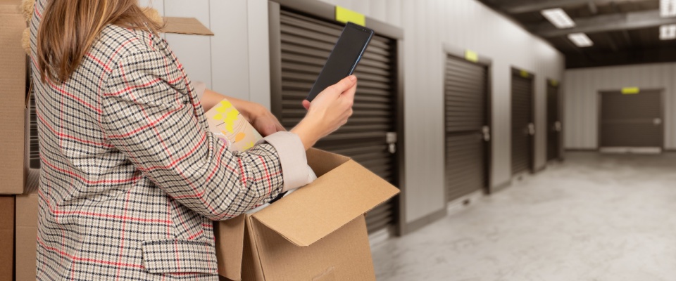 Woman unpacking a cardboard box and using a mobile phone to look for additional insurance coverage, while standing in a hallway lined with storage unit doors.