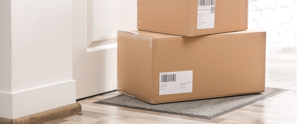 Two cardboard delivery boxes stacked on a doormat outside a white front door, illustrating how to protect your home while on vacation by pausing package deliveries or asking a friend to pick up packages.