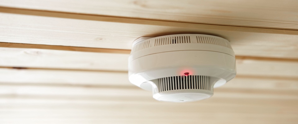 Smoke detector with a glowing red indicator light mounted on a wooden ceiling, to be checked before going on vacation in order to  protect the rental from fire hazards.