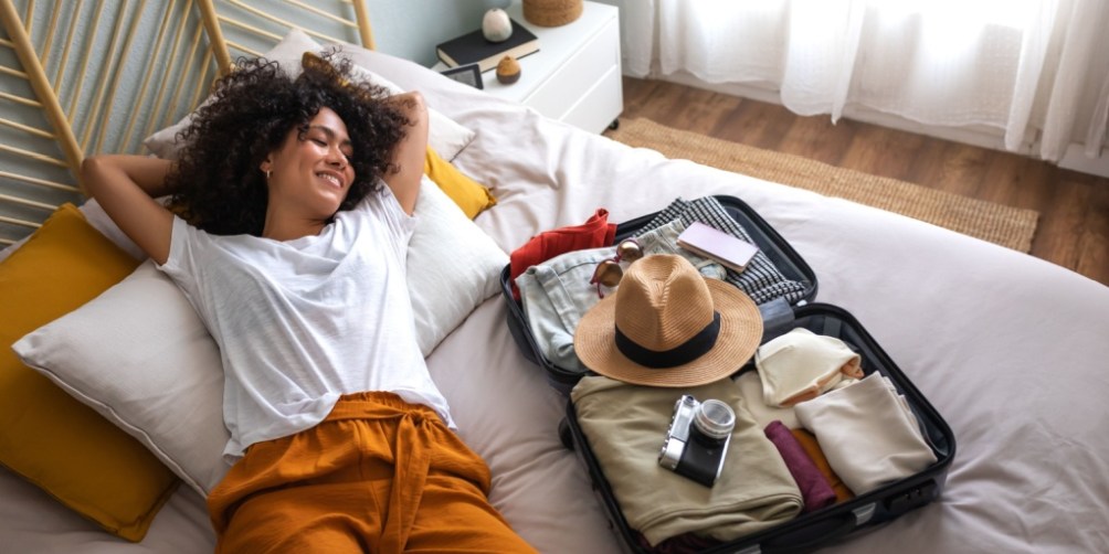 Woman relaxing on a bed in a bright, cozy bedroom setting, next to an open suitcase packed with clothes, a hat, sunglasses, and a camera, after learning how to protect her home while on vacation.