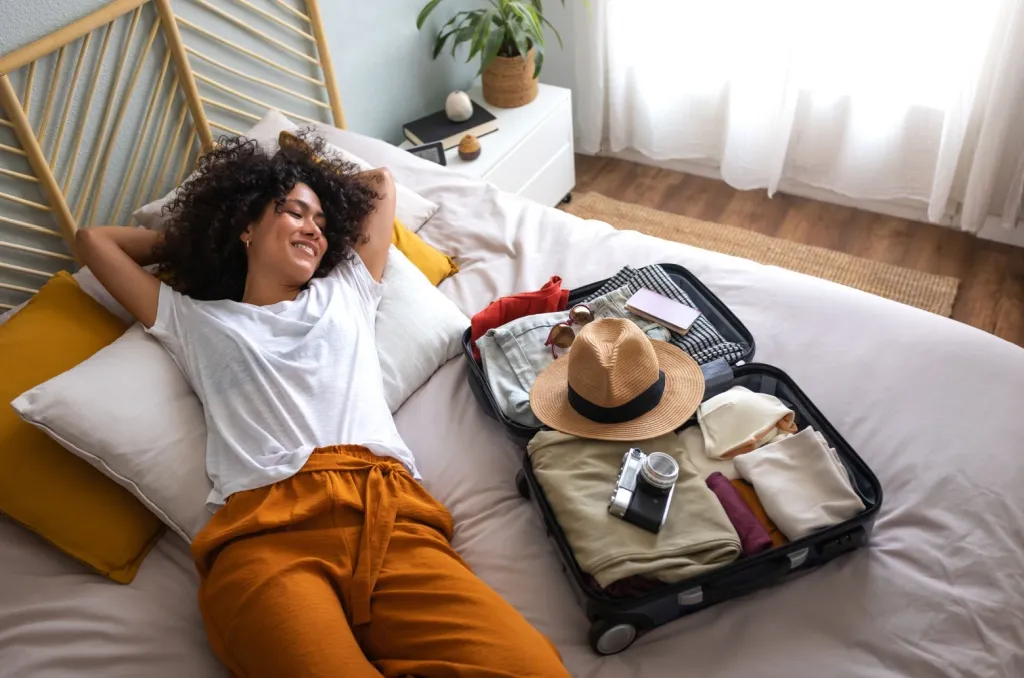 Woman relaxing on a bed in a bright, cozy bedroom setting, next to an open suitcase packed with clothes, a hat, sunglasses, and a camera, after learning how to protect her home while on vacation.