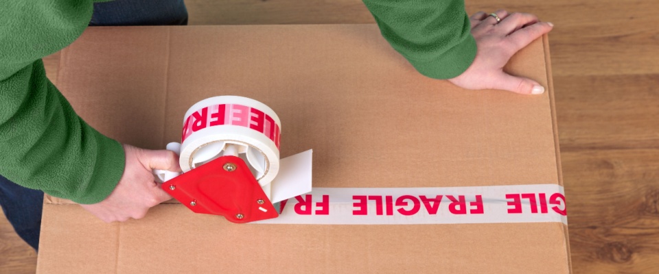 Person sealing a cardboard box labeled "fragile" with packing tape, illustrating what renters insurance doesn't cover during moving, such as items damage or losses that occur while packing and transporting belongings.