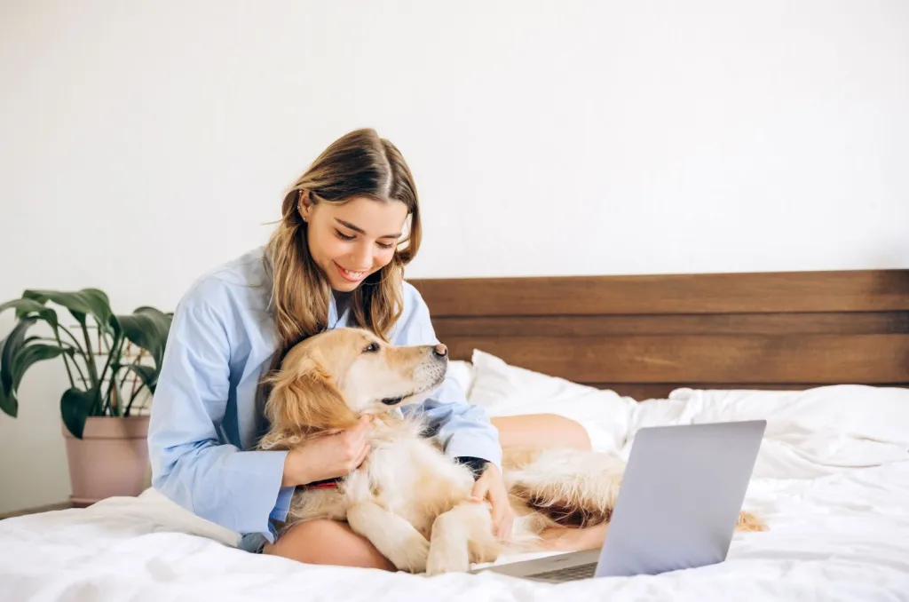 Woman sitting on a bed in a bright bedroom, gently hugging her golden retriever next to an open laptop, illustrating a common scenario for pet owners wondering if renters insurance covers pet damage.