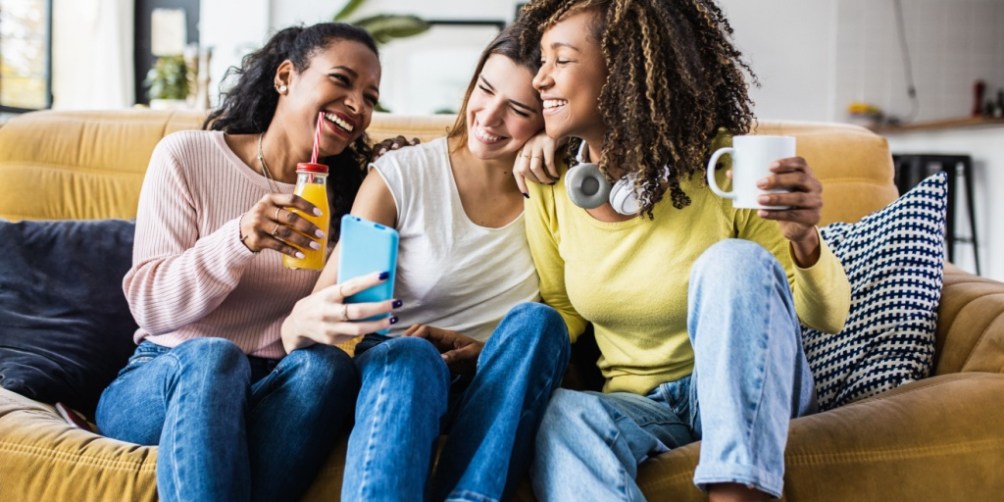 Three college students sitting together on a cozy sofa, holding drinks and a smartphone, while discussing the importance of student renters insurance for protecting personal belongings and electronics in a shared apartment.