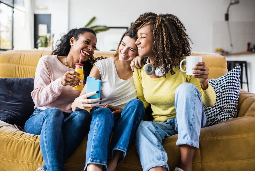 Three college students sitting together on a cozy sofa, holding drinks and a smartphone, while discussing the importance of student renters insurance for protecting personal belongings and electronics in a shared apartment.