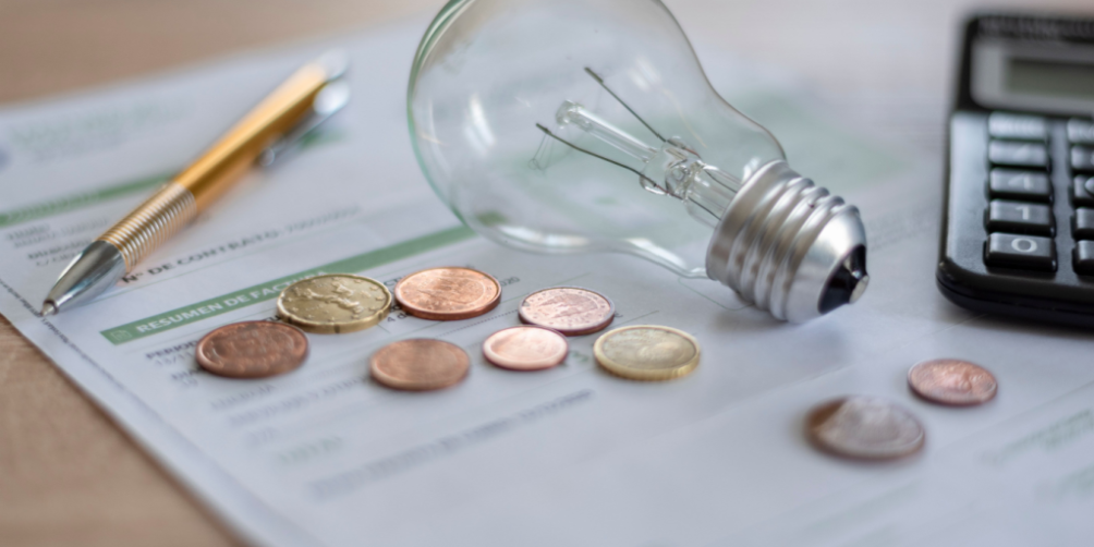 Electricity bills covered by several coins, a light bulb, calculator, and pen on a desk.