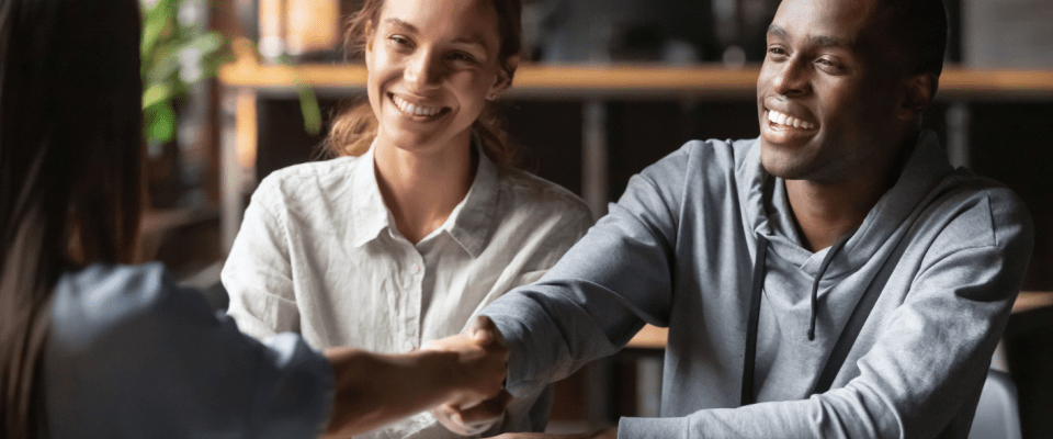 A happy young couple sitting at a desk and shaking hands with their landlord or property manager after agreeing on the terms of renters insurance required by the landlord.