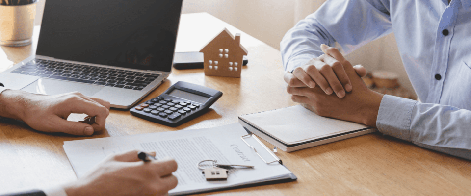 Two people reviewing a contract and discussing financial calculations, with a laptop, calculator, and a small house model on the table, emphasizing important details about what renters insurance doesn't cover.