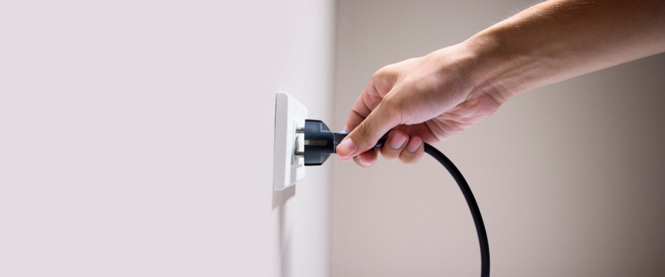 Close-up of a hand inserting a black electrical cord into an outlet on a white wall, highlighting the importance of handling plugs properly as an electrical safety tip.