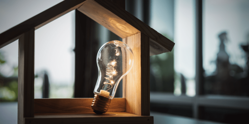 Wooden house-shaped frame with a glowing light bulb inside, symbolizing home utilities and how to set up electricity in an apartment.