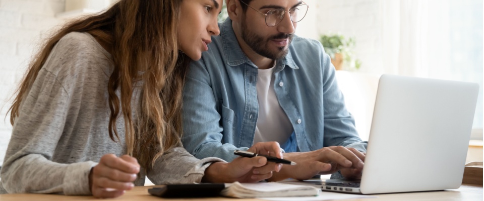 A young man and woman looking at a laptop screen together while sitting at a table with papers and a calculator, paying rent, utilities, or renters insurance, showcasing how various factors may or may not affect credit score.