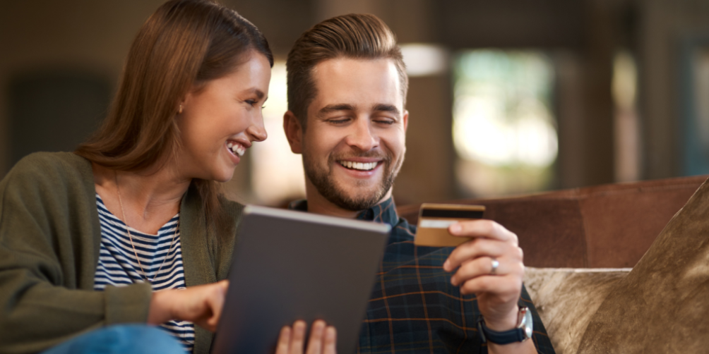 Happy young couple sitting on a couch, holding a credit card, and using a tablet to find out whether renters insurance affect credit score and financial planning.