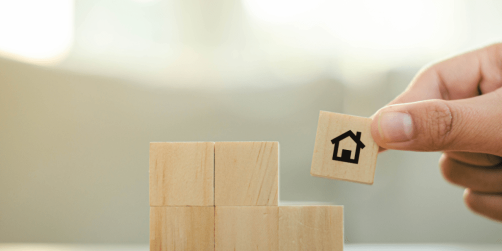 Wooden blocks stacked to form a foundation with a hand placing a block featuring a house icon on top, symbolizing coverage concepts and illustrating the topic 'what renters insurance doesn't cover.’
