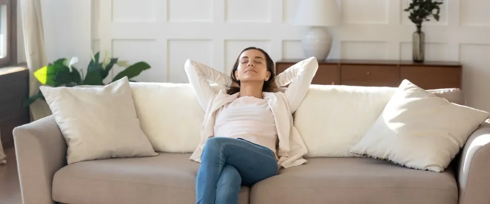 Woman relaxing on a modern beige sofa in a bright, airy living room filled with plants and natural light, illustrating the benefits of clean air and the impact of renewable resources on a healthy home environment.