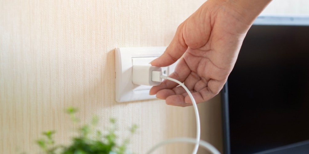Hand unplugging a phone charger from a wall outlet next to a TV and houseplant, illustrating how unplugging appliances saves electricity by preventing phantom energy use.