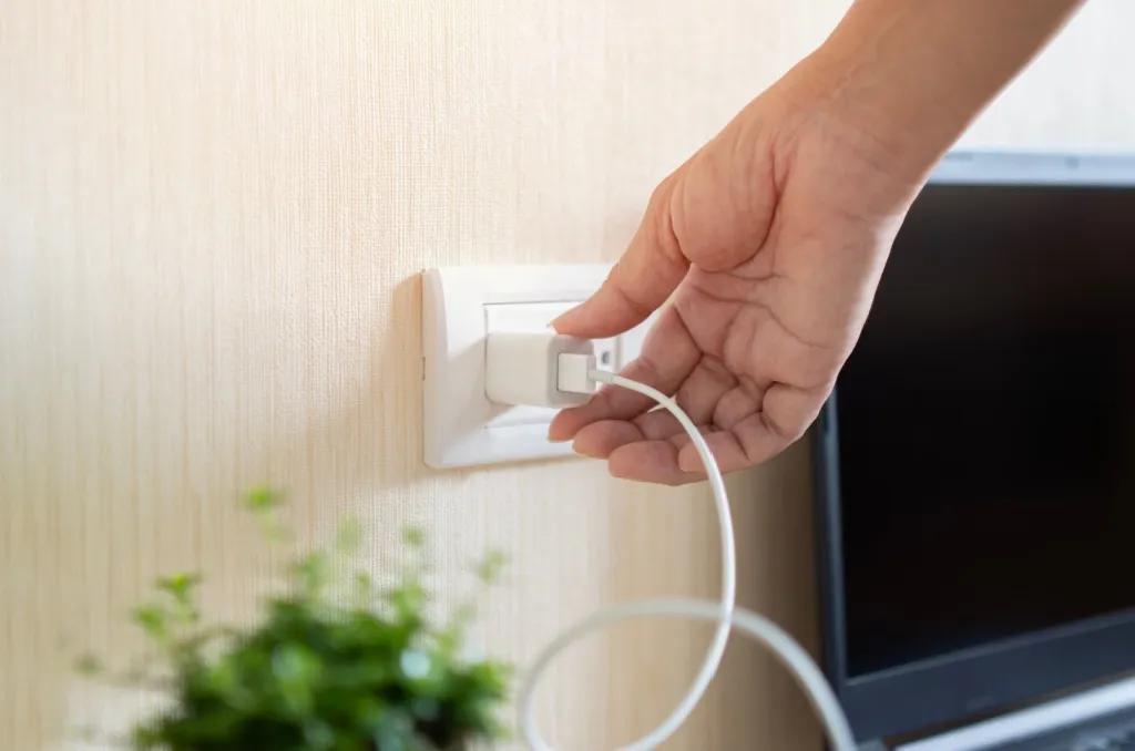 Hand unplugging a phone charger from a wall outlet next to a TV and houseplant, illustrating how unplugging appliances saves electricity by preventing phantom energy use.