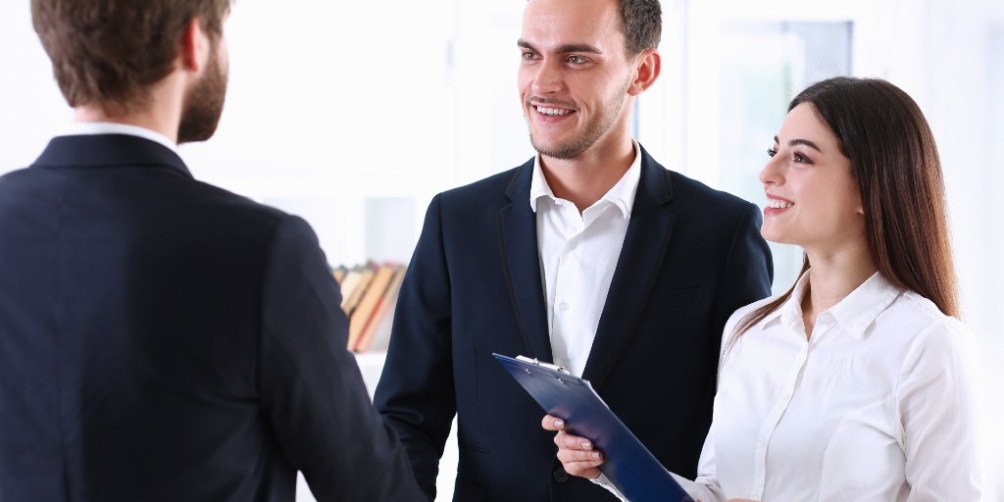 Two men elegantly dressed shaking hands while a woman holding a clipboard smiles, discussing terms involving a guarantor or a cosigner.