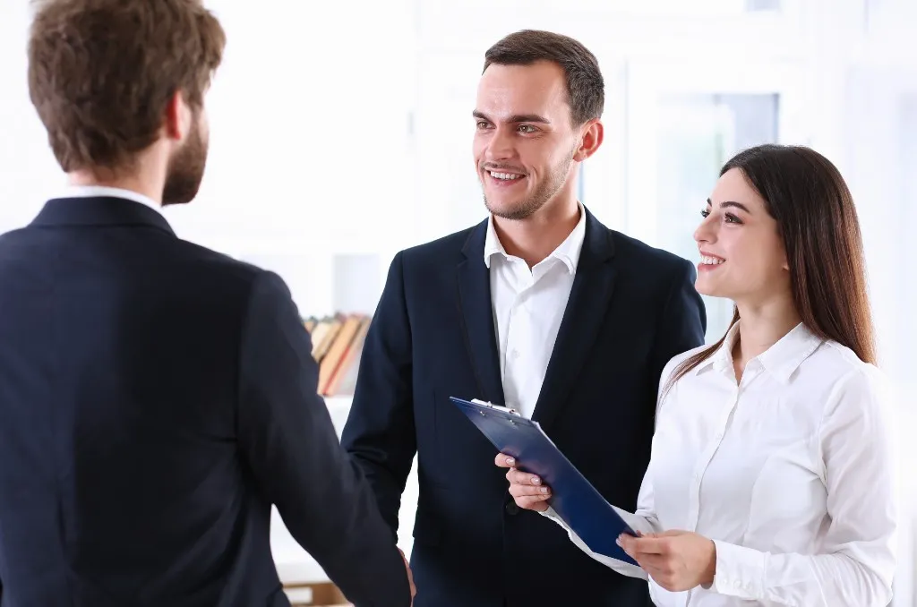 Two men elegantly dressed shaking hands while a woman holding a clipboard smiles, discussing terms involving a guarantor or a cosigner.
