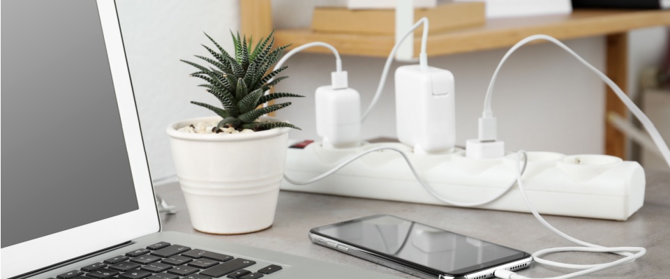 Power strip on a desk with multiple phone chargers, a laptop, and a smartphone, visually demonstrating the importance of unplugging appliances to save electricity.
